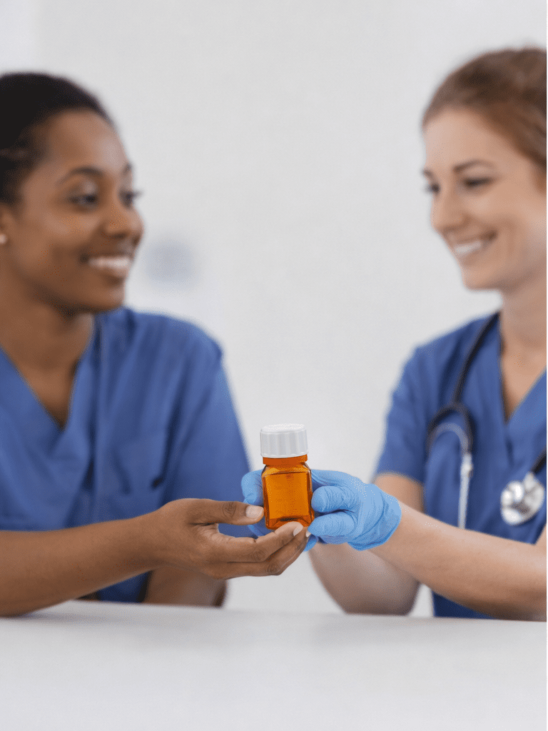 January_blog_1 - The LiquiMedLock Blog Two female nurses in blue scrubs sit and smile in a bright clinical setting; one nurse is holding a small orange square liquid medicine bottle with a white child-resistant and tamper evident cap in the foreground.